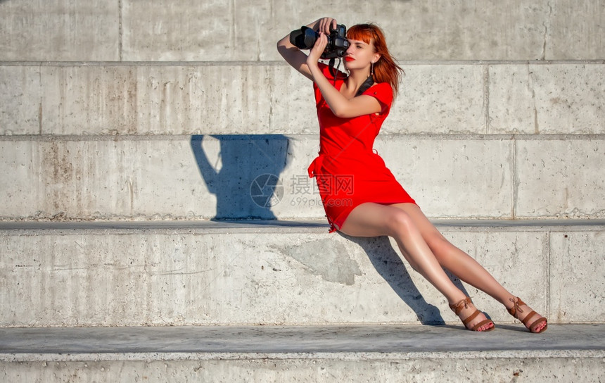 穿着亮红礼服的年轻美女摄影师专注于一个配有大型数码相机的模特图片