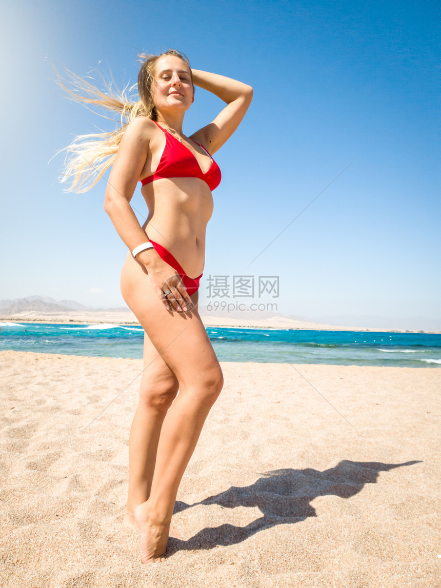 美丽的瘦女人长发穿着感红色比基尼假扮在海滩上浪和蓝天空面对海浪和蓝天空美丽的瘦年轻女人长发穿着感红色比基尼在海滩上浪和蓝天图片