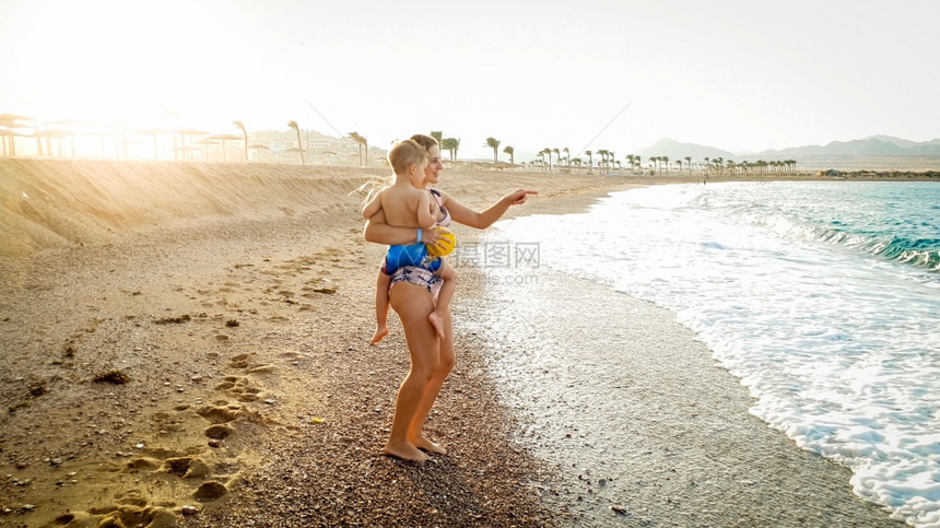 年轻母亲在沙海滨抱三岁小儿子看着平静的海浪和日落天空图片