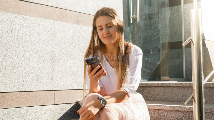 美丽的微笑年轻女孩坐在石头的楼梯上在智能手机浏览社交媒体图片