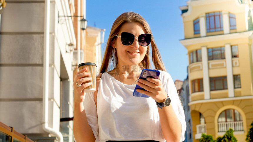 带着一杯咖啡在街上行走并使用智能手机的美丽微笑年轻女子肖像图片