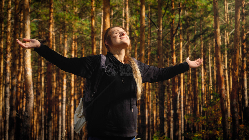 美丽的微笑女游客肖像背包伸展的手在松树林中仰望明亮的太阳自由概念和与然谐相处的概念背包伸展的手在松树林中仰望明亮的太阳自由以及与图片