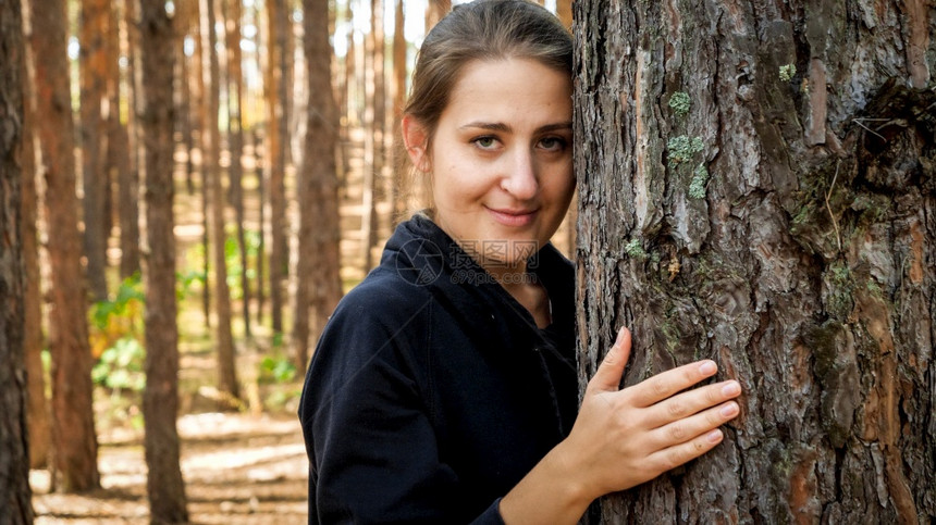 微笑的年轻女子在森林里拥抱松树并秘密地看着爱生态保护和与自然谐相处的概念微笑年轻女子在森林里拥抱松树秘密地看着图片