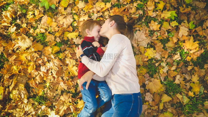 美丽的年轻女子在秋天公园的树叶上亲吻她小儿子图片