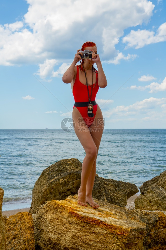 一个穿着紧闭红色泳衣的年轻女孩带着一个反光相机和测量仪站在岩石海滩上拍照图片