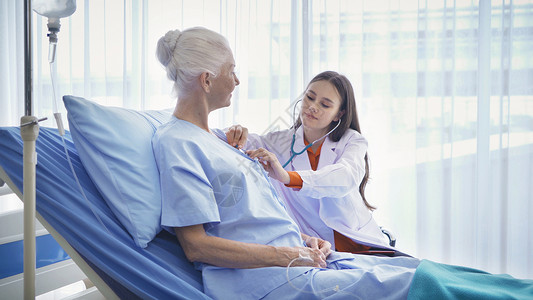 治未病快乐的女医生通过听诊器检查生病的老年女病人的身体或心脏她们躺在医院病房的床上接受医疗和治背景