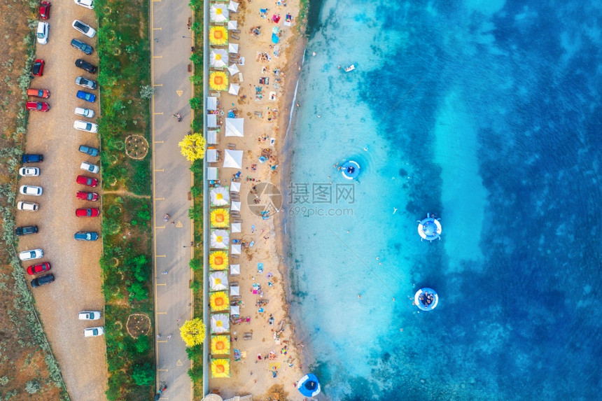 沙滩的空中景象有多彩的雨伞在海上游泳的人有透明蓝水游泳环绿树和汽车在夏季日落的赛道上图片