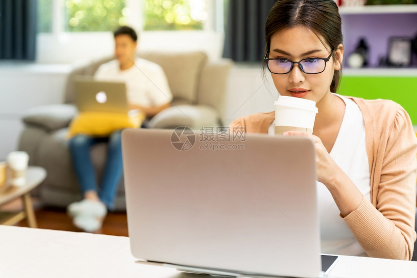 亚洲工作妇女使用电脑笔记本电脑送咖啡杯回家工作在有社交距离的客厅里工作在城市被封锁时背景情侣图片