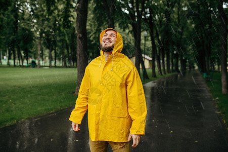 穿着雨衣的男子享受夏季公园的雨穿着披风的男子在步行路上快乐巷子里的湿天气穿雨衣的男子享受夏季公园的雨背景图片