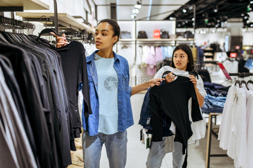 两个女孩穿着衣服在装店的架上穿服妇女在时装店商购物看服装的购物者女孩穿着衣服在装店的架上穿服图片