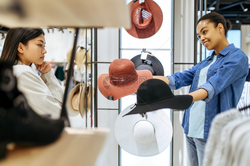 在服装店中选择帽子的两个女孩在时装店商购物的妇女在衣架上看服装的购物妇女在服装店中选择帽子的两个女孩图片