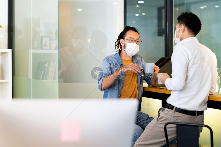 两名办公室工作人员在喝咖啡时交谈而在新正常状态中与社会远程执业办公室交谈他们戴着面罩减少作为新正常生活方式的新冠19日冕感染的风图片