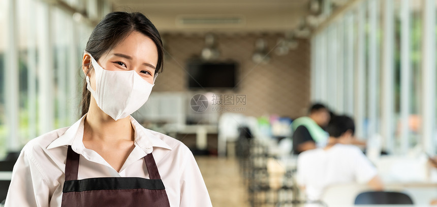 具有吸引力的亚洲女服务员面带顾客背景的孔坐在室内餐厅的社会距离桌子上图片