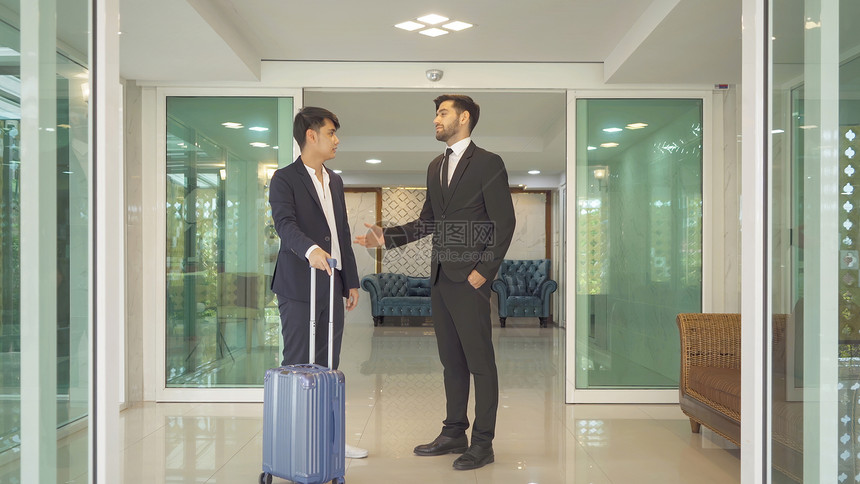 带着行李到大厅走廊和在酒店与同事交谈的亚洲商人微笑着团队合作伙伴在出差旅行中进了讨论图片