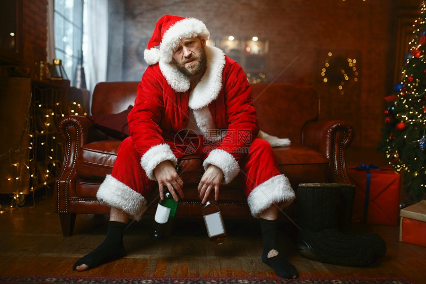 坏的圣誕老人庆祝诞节在沙发上用一瓶酒精庆祝圣诞节不健康的生活方式在节日服装新年和酗酒中留胡子的男用瓶庆祝圣诞节图片