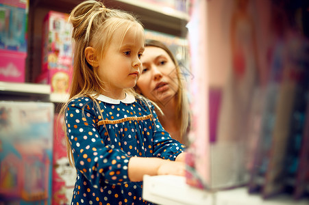 妈和可爱的女儿在玩具店展台附近快乐的童年家庭在商店买东西妈和小女孩在玩具店里买一个娃玩具店背景图片