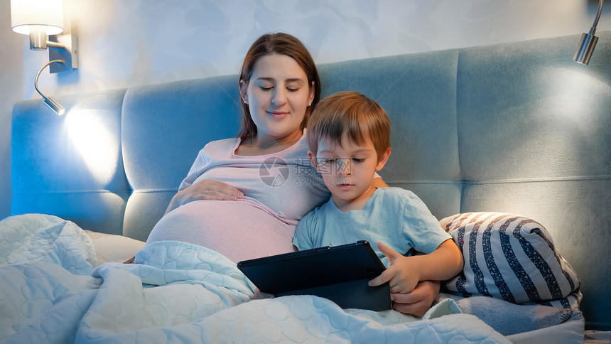 快乐有怀孕母亲的男孩在睡觉前晚上平板电脑看卡通快乐男孩在睡觉前晚上平板电脑看卡通图片