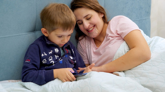 幼儿男孩与年轻母亲睡在床上平板电脑玩游戏图片