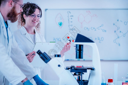 年轻科学家在实验室穿白外套的显微镜前笑和站立董事会图片