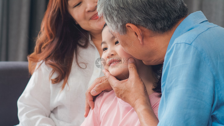亚洲祖父母在家里亲吻孙女的脸颊高龄人老一代祖父和母利用家庭时间放松在客厅概念中年轻女孩躺在沙发上图片