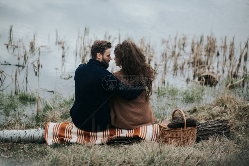 2个天主教情人坐在湖边的毯子上年轻夫妇在秋天的户外拥抱一个留胡子的男人和卷毛女恋爱情节爱与家庭的概念背面看两个坐在湖边毯子上的天图片