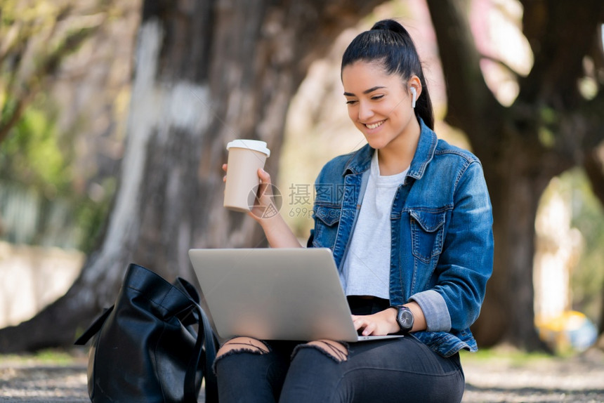 青年妇女坐在户外时使用笔记本电脑和喝咖啡的肖像图片
