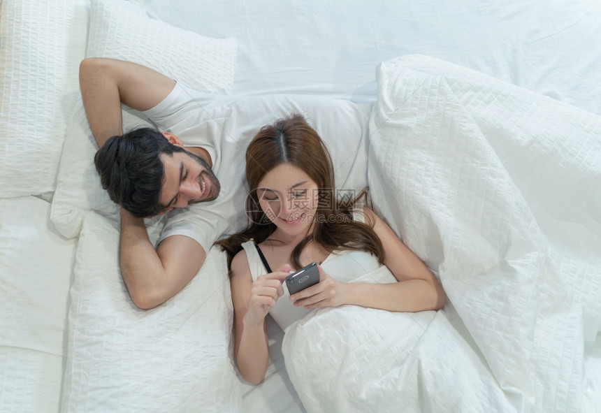 一对夫妇家庭中东青年快乐和亚洲女在社交媒体互联网上使用智能手机的人和在家中卧室睡觉的人早期技术中的生活方式图片