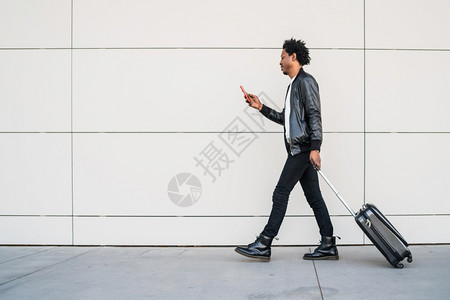 Afro旅游男子在街上户外行走时使用其移动电话和携带手提箱的肖像图片