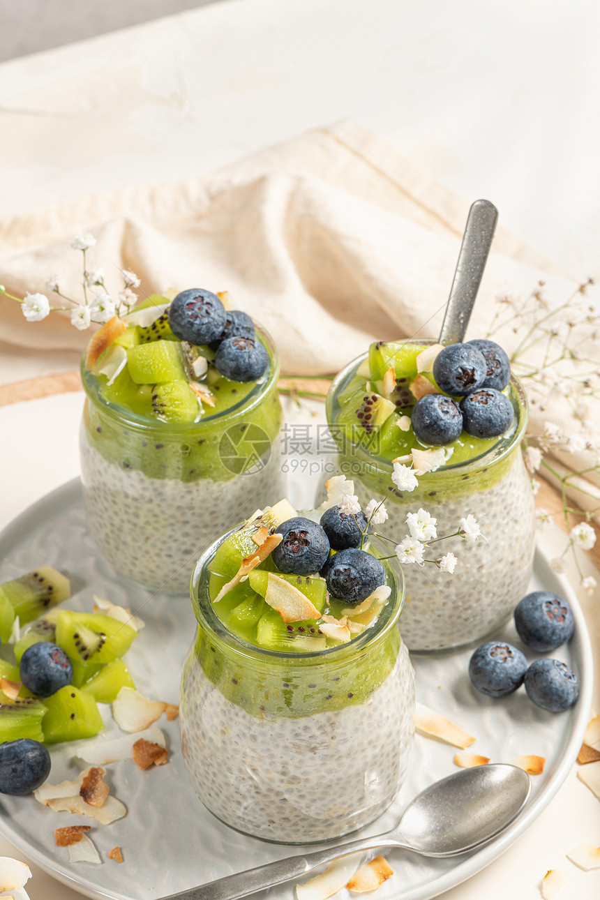健康早餐辣椒布丁配有kiwi蓝莓和椰子片三块放在白桌上的玻璃罐中图片