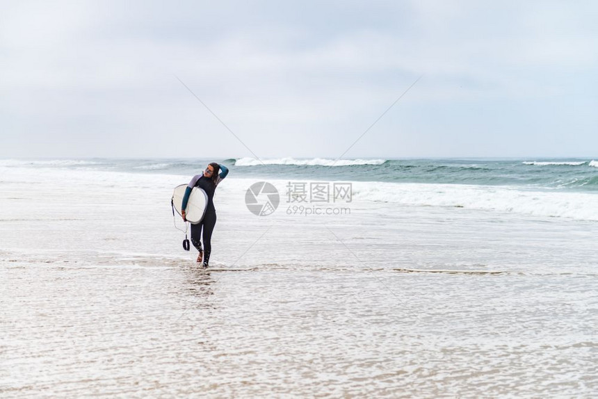 年轻女冲浪者穿着泳衣手臂下握着冲浪板早上后在海滩散步图片