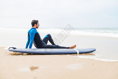 年轻男冲浪者穿着泳衣早上冲浪课后坐在海滩上的冲浪板旁图片