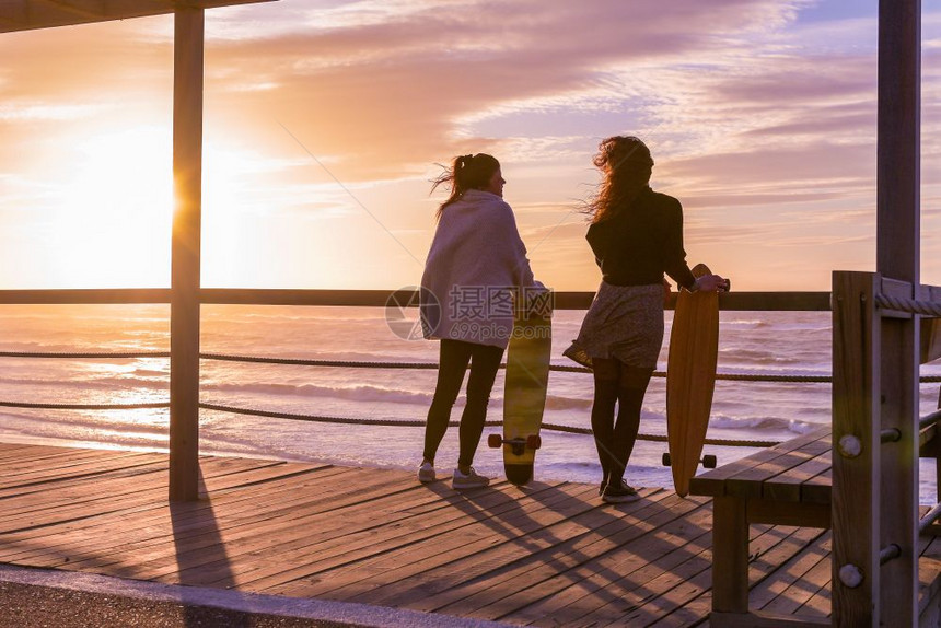 日落时两个女朋友在海滩附近与滑板聊天图片