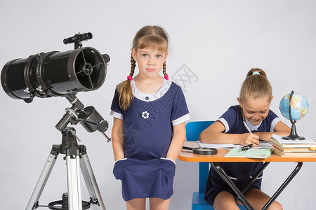 一个女孩站在望远镜旁手放在口袋里另一个女孩坐在桌子上写字图片