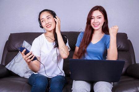 两名年轻妇女使用笔记本电脑在家中沙发上用便器耳机收听音乐图片