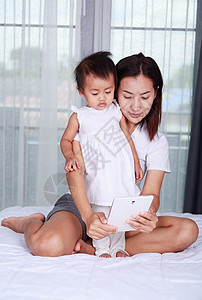 母亲和婴儿正在家里的床上玩耍和阅读平板电脑图片