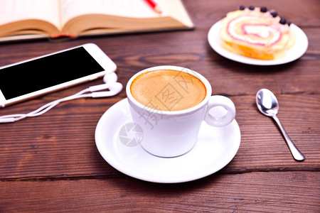 棕色木背景的咖啡杯背在带耳机和一本公开书的智能手机后面图片