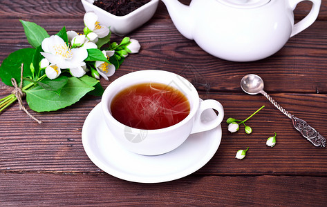 白杯中的热黑茶与一个碟子背后白色茶壶和一束花朵的茉莉束图片