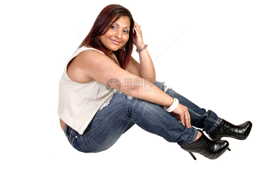 一个微笑的西班牙年轻女人穿着破烂的牛仔裤穿着黄油和靴子坐在地板上做白底衣图片