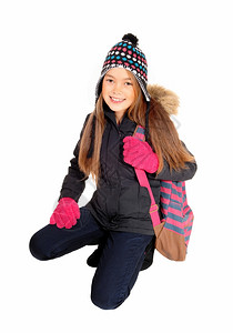 一个美丽的小女孩带着帽子和手套背包在她的肩膀上跪下与白种背景隔绝图片