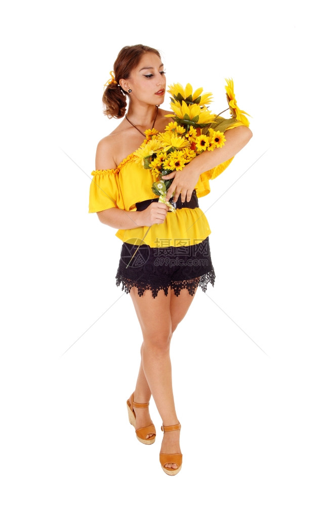 穿着黄色上衣的迷人年轻女子拿着一堆向日葵看着他们站为白背景而孤立图片