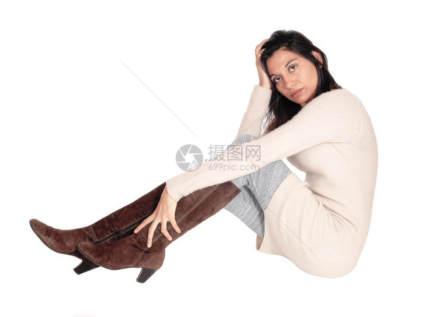 一位美丽的西班牙年轻女穿着一件针织的裙子穿着棕色的靴子坐在地板上面部她手孤立白色背景上图片