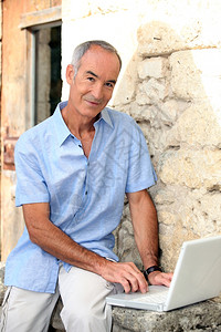 灰色头发男子用笔记本电脑坐在石墙边图片