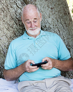 坐在树下户外的老人在他智能手机上发短信图片