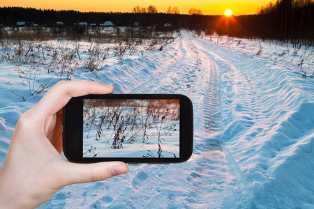 旅行概念旅游者用智能手机拍摄田间蓝色冬季雪堤的日落景象图片