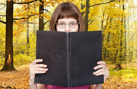 带着眼镜的女孩阅读大书空白的封面和黄色秋林背景图片