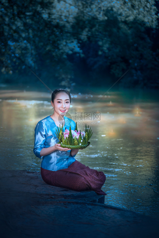 身着传统亚洲服饰的美丽女手持由香蕉叶和花朵制成的容器坐在河边LoyKrathong传统节图片