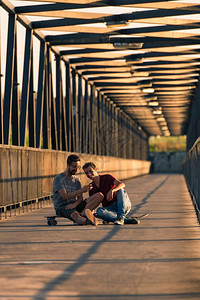 年轻的滑板运动员坐在外面的板子上坐在河边的行人桥上发短信图片