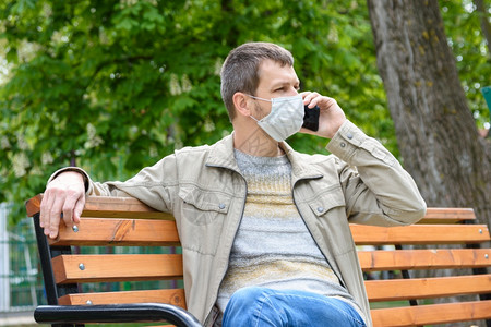 一个戴保护面罩的人穿过公园蹲在一张长椅上讲电话图片