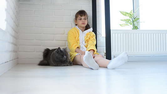 一个穿着黄色浴袍的不开心女孩坐在房间的角落一只深灰色家猫坐在附近背景图片