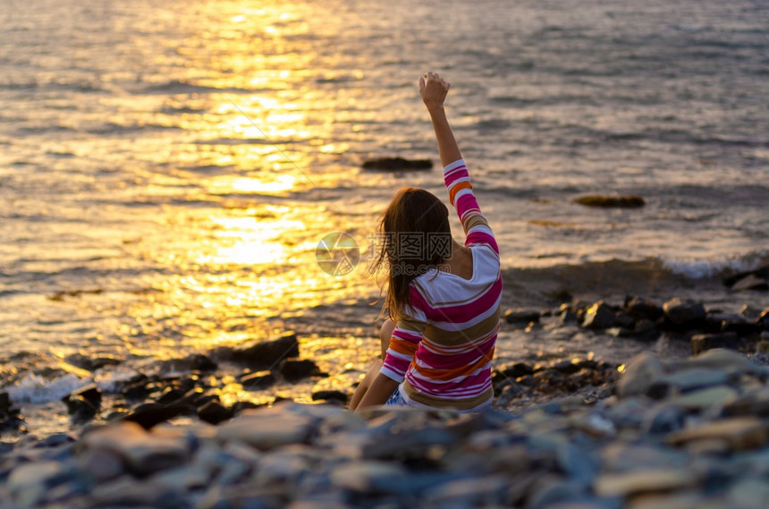一个女孩坐在海边向水里扔石头举起手来晚上日落图片